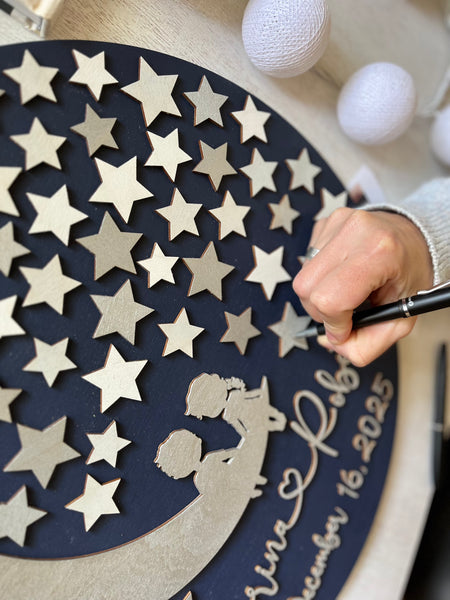 A hand shown to sign a decorative wall art wedding guest book with stars and a silhouette of a couple, featuring personalized text.