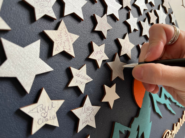 Hand using a pen to sign star-shaped pieces on a 3D wood wedding guest book alternative personalized with the names f the bride and groom