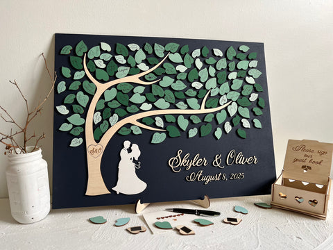 Magnetic guest book alternative for wedding made on a rectangular magnetic board with a tree and magnetic leaves for the guests to sign. The board is navy blue and the leaves are in various shades of green and sage green. The guest book is displayed on a stand (included in the purchase). There is a box to store the leaves, the top of the box is engraved with the text "Please sign our guest book". 