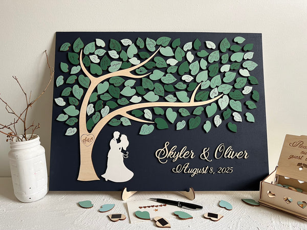 magnetic guest book alternative for wedding made on a rectangular magnetic board with a tree and magnetic leaves for the guests to sign. The board is navy blue and the leaves are in various shades of green and sage green. The guest book is displayed on a stand (included in the purchase). There is a box to store the leaves, the top of the box is engraved with the text "Please sign our guest book". 