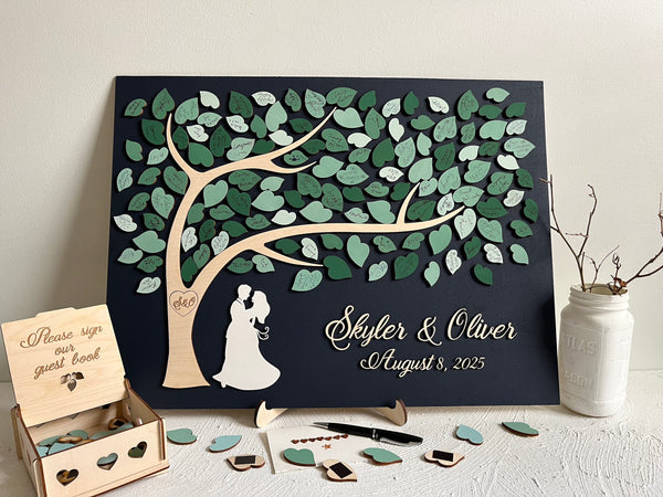 Magnetic guest book alternative for wedding made on a rectangular magnetic board with a tree and magnetic leaves for the guests to sign. The board is navy blue and the leaves are in various shades of green and sage green. The guest book is displayed on a stand (included in the purchase). There is a box to store the leaves, the top of the box is engraved with the text "Please sign our guest book". 