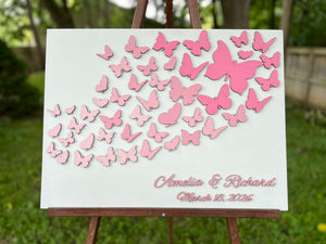 Butterfly wedding guest book with personalized names and date on a wood white board shown in an outdoors setting. The butterflies as well as the personalized text are all in pink but can be customized to reflect your preference. 