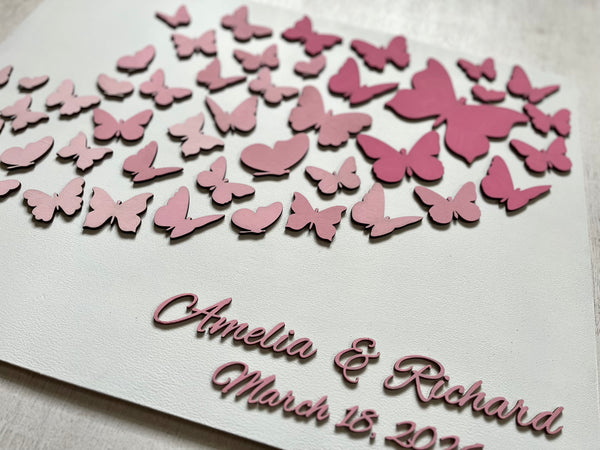 Butterfly wedding guest book with personalized names and date on a wood white board, shown in a closeup. The butterflies as well as the personalized text are all in pink but can be customized to reflect your preference. 