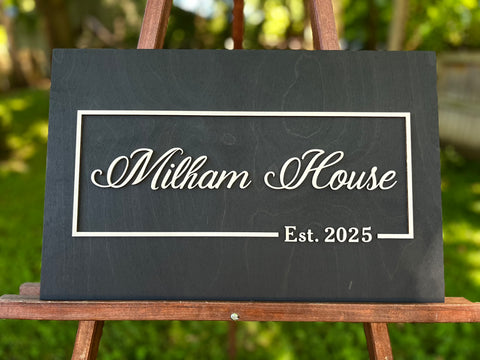 Black sign with 'Family Last name and House Est. 2025' on a wooden stand outdoors, Christmas gift