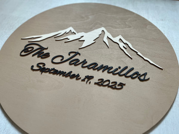 Round wooden plaque with mountain design and 3D  text on a tan /beige background