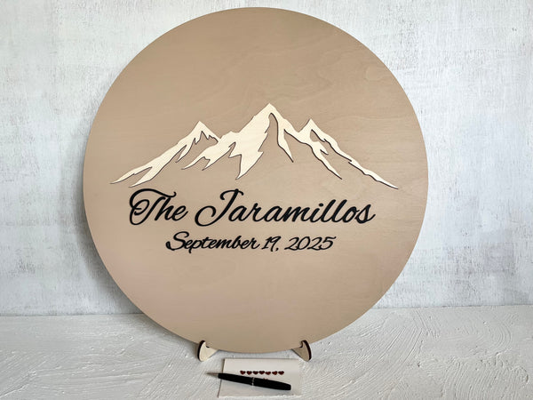 Round wooden sign with mountain design and 3D text on a light background- used as a wedding guest book alternative for guest signatures