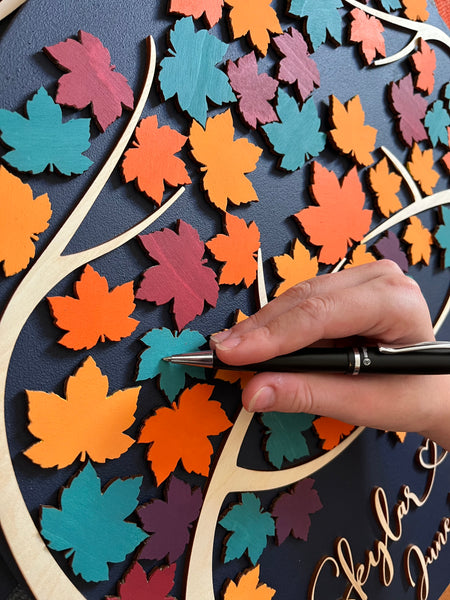 Hand holding a pen over a wedding guest book with  colorful maple leaf patterns on a dark navy background.