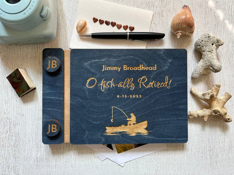Personalized retirement guest book with fishing design and  the engraved text'O fish-ally retired!' text on a wooden surface. Ideal for a retirement celebration with a fishing theme, personalized for the person celebrating the birthday or retirement. The guest book cover is custom engraved with a fishing boat and a fisherman and has custom engraved buttons with the initials of the retiree.
