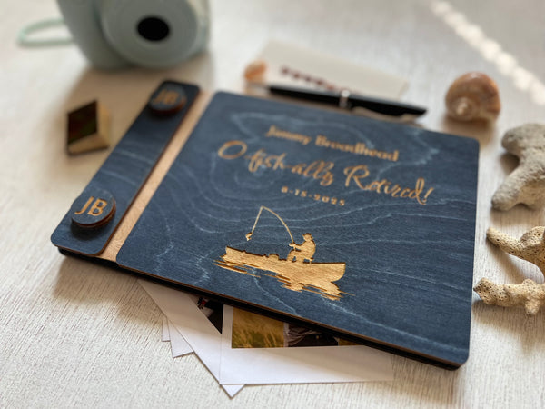 Personalized retirement guest book with fishing design and  the engraved text'O fish-ally retired!' text on a wooden surface. Ideal for a retirement celebration with a fishing theme, personalized for the person celebrating the birthday or retirement. The guest book cover is custom engraved with a fishing boat and a fisherman and has custom engraved buttons with the initials of the retiree.