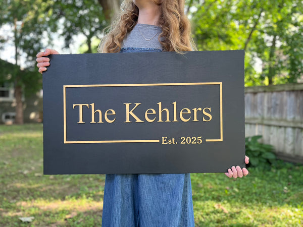 Person holding a sign with persoanlized last name and established date in an outdoor setting