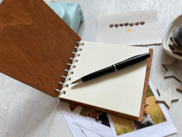 Open notebook with a pen on top, surrounded by photos and stationery items on a textured surface.