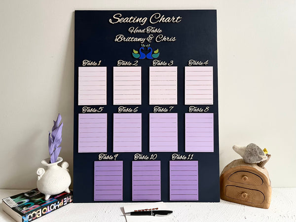 Seating chart for a wedding with purple placeholders on a dark navy wood board, personalized with the names of the bride and groom and a pair of peacock silhouettes.