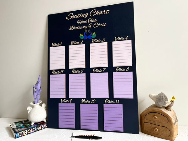 Seating arrangement sign for a wedding with purple placeholders on a dark navy wood board, personalized with the names of the bride and groom and a pair of peacock silhouettes.