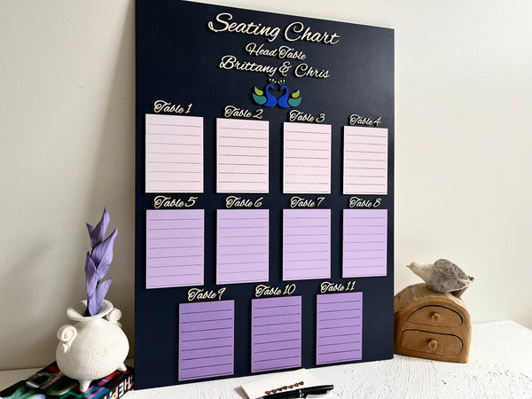 Seating chart for a wedding with purple placeholders on a black board, placed on a surface with decorative items.