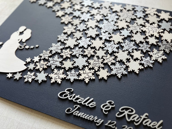 Wedding guest book wall art with snowflakes to sign, personalized names, wedding date and silhouettes of a couple on a navy blue background. The snowflakes on the guest book are signed by wedding guests