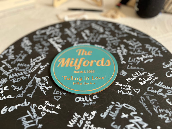 Vinyl music record lookalike sign used as a wedding guest book with personalized names, date and special song title and artist engraved in the middle. The wedding guests sign the record 