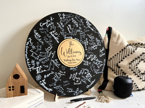 Round black wooden music record used as a wedding guest book for guest signatures. Personalized with the last name of the couple, a wedding date as well as a favourite song and artist, engraved in the middle of the record, to mimic a musical vinyl record.