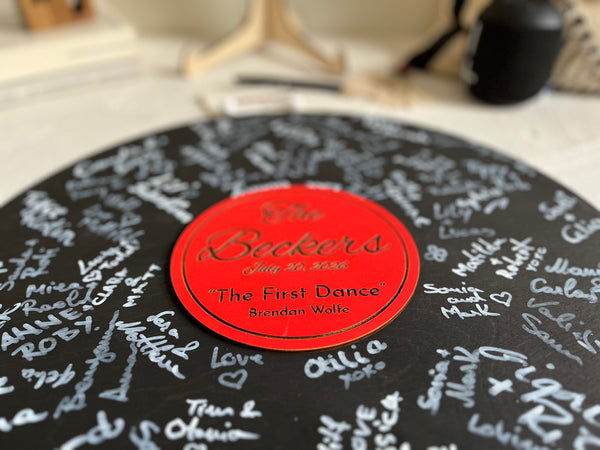 Vinyl music record lookalike sign used as a wedding guest book with personalized names, date and special song title and artist engraved in the middle. The wedding guests sign the record 