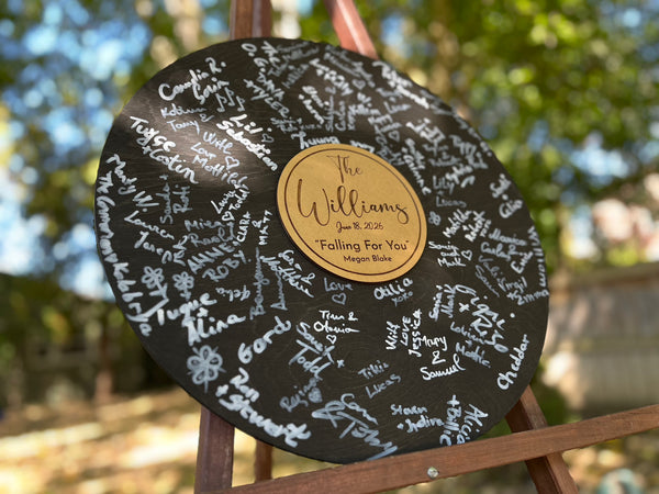 Round black wooden music record used as a wedding guest book for guest signatures. Personalized with the last name of the couple, a wedding date as well as a favourite song and artist, engraved in the middle of the record, to mimic a musical vinyl record.