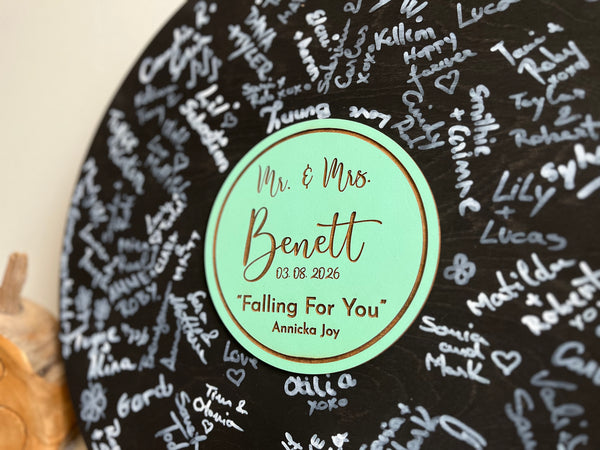 Vinyl music record lookalike sign used as a wedding guest book with personalized names, date and special song title and artist engraved in the middle. The wedding guests sign the record 
