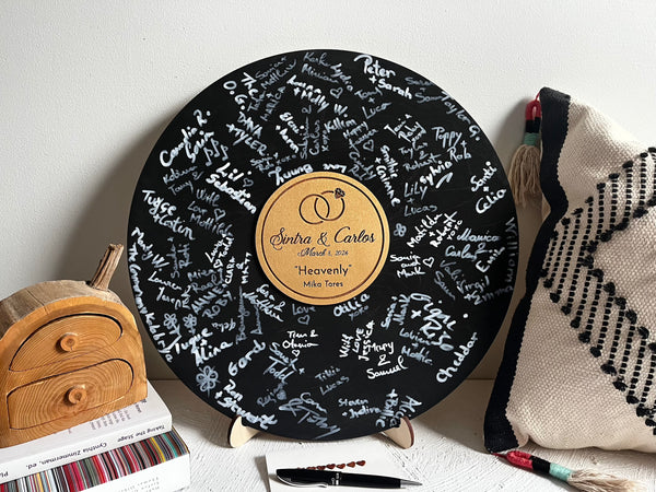 Black round vinyl record looking guest book made of wood for guest signatures, personalized with engraved text. The board is shown after the guests have signed it