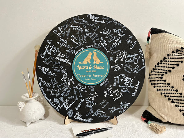 Decorative vinyl record with engraved names and a stand, surrounded by decorative items on a light surface. Used as an alternative weddng guest book to gather guests' signatures