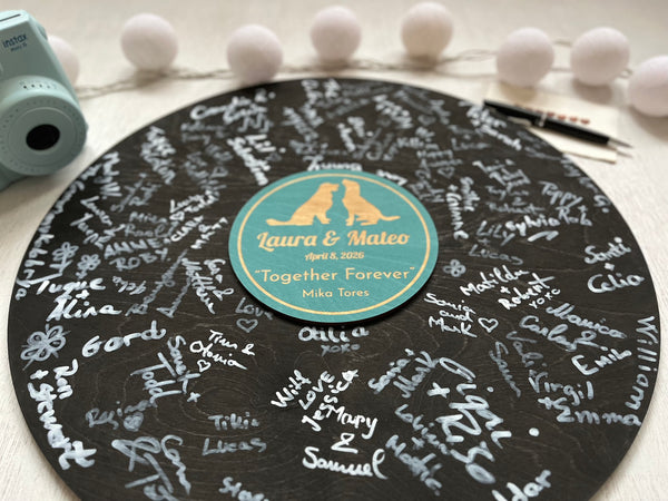 Decorative vinyl record with engraved names and a stand, surrounded by decorative items on a light surface. Used as an alternative weddng guest book to gather guests' signatures