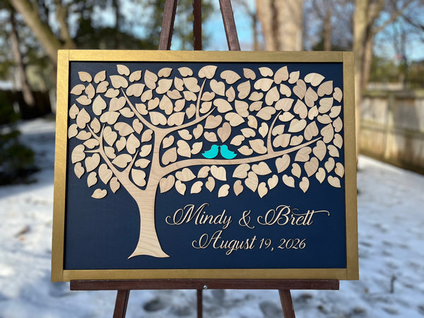 Framed guest book with a tree design, two blue birds, and personalized text on a navy background shown in a snowy setting.