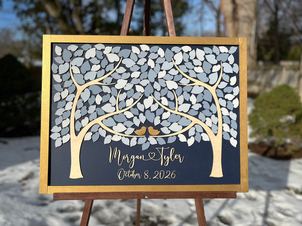 A personalized wooden wedding tree sign with grey and white painted leaves, gold  birds, and personalized script text with the names of the bride and groom as well as wedding date displayed on an easel outdoors in a snowy setting.