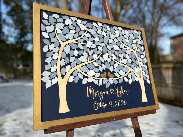 A personalized wooden wedding guest book sign mounted on an easel outdoors. The sign features a gold tree design with white and light grey leaf cutouts, two small gold birds, and gold script text featuring the names of the bride and groom and their wedding date, all  on a dark navy background.