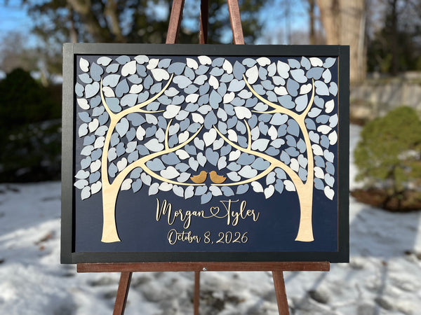 A framed personalized wedding sign displayed on an easel outdoors. The sign features a dark navy background with a stylized tree made of white and gray leaf shapes, two small gold birds, and script text personalized with the names and wedding or anniversary date