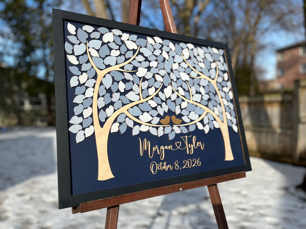 A framed personalized wedding sign displayed on an easel outdoors. The sign features a dark navy background with a stylized tree made of white and gray leaf shapes, two small gold birds, and script text personalized with the names and wedding or anniversary date