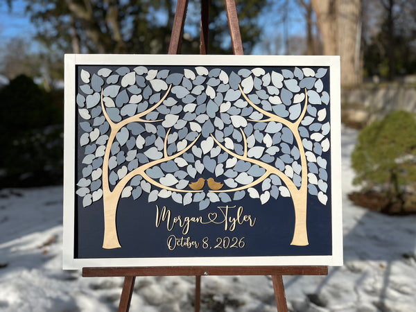 A framed personalized wedding sign displayed on an easel outdoors. The sign features a dark navy background with a stylized tree made of white and gray leaf shapes, two small gold birds, and script text personalized with the names and wedding or anniversary date