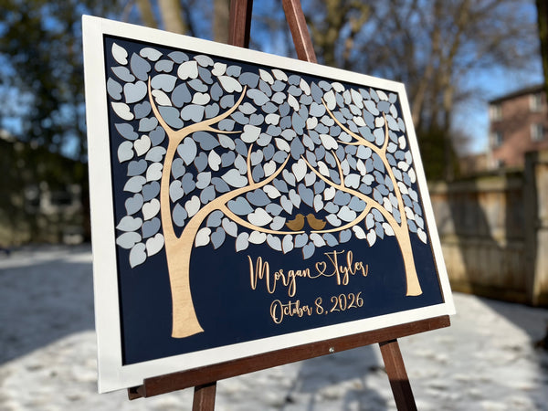 A framed personalized wedding sign displayed on an easel outdoors. The sign features a dark navy background with a stylized tree made of white and gray leaf shapes, two small gold birds, and script text personalized with the names and wedding or anniversary date