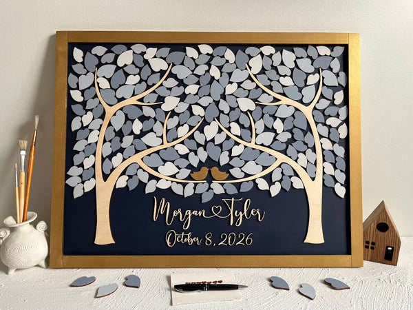 A framed personalized wooden tree wedding sign with layered gray and white leaves, two small gold birds, and custom names and the wedding or anniversary date on a dark navy background. The piece is styled on a white surface.