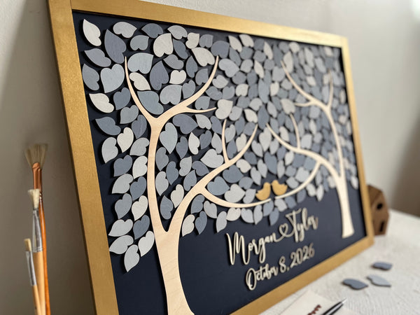 A framed personalized tree-of-life wall art piece with layered gray and white leaf shapes, a gold frame, and custom personalized text on a dark background, photographed indoors on a tabletop.