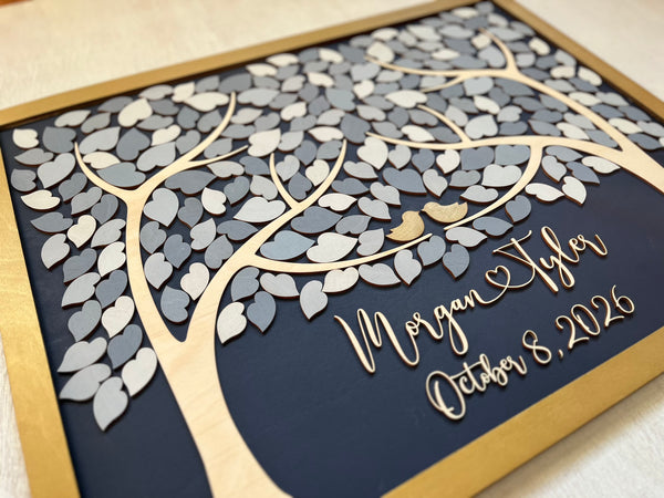 A detail shown of a rectangular wooden personalized family tree wall art piece with layered laser-cut leaves in light and dark gray, a gold-toned frame, and custom script text with the names and wedding date on a dark navy background.
