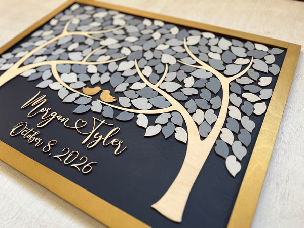 A detail shown of a rectangular wooden personalized family tree wall art piece with layered laser-cut leaves in light and dark gray, a gold-toned frame, and custom script text with the names and wedding date on a dark navy background.