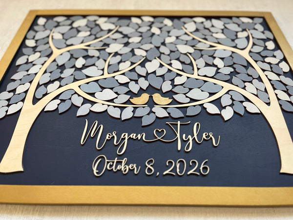 A detail shown of a rectangular wooden personalized family tree wall art piece with layered laser-cut leaves in light and dark gray, a gold-toned frame, and custom script text with the names and wedding date on a dark navy background.