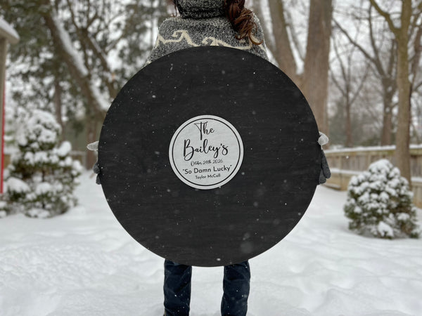Person holding a large black disc with a personalized label in a snowy outdoor setting