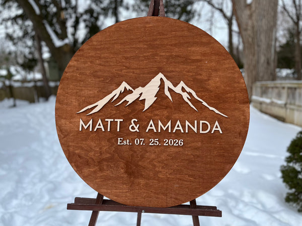Wooden plaque with mountain design personalized names and established date  in a snowy outdoor setting.
