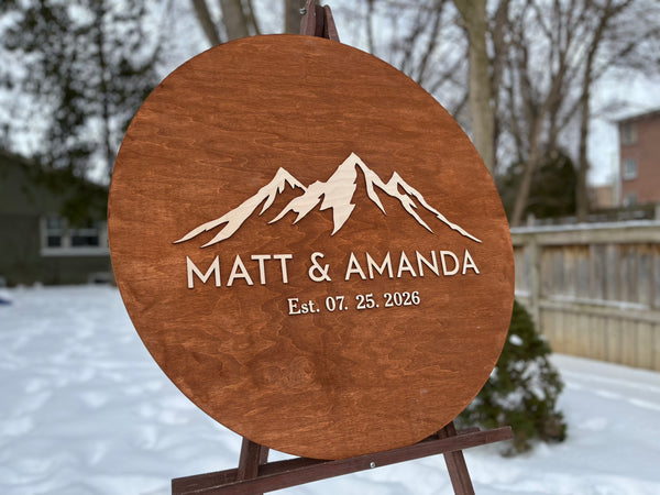 Wooden plaque with mountain design personalized names and established date  in a snowy outdoor setting.