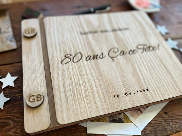A personalized wooden keepsake album or photo box with a light natural wood grain cover, engraved with '80 ans Ca ce Fete!' and a date, photographed on a wooden tabletop with scattered star-shaped paper and photos around it.