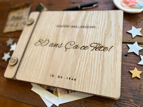 A personalized wooden keepsake album or notebook cover laid flat on a rustic wooden table, with the engraved text  '80 ans Ça ce Fête!', and a personalized name and year