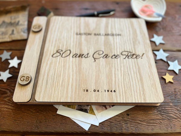 A personalized wooden keepsake album or notebook cover laid flat on a rustic wooden table, with the engraved text  '80 ans Ça ce Fête!', and a personalized name and year