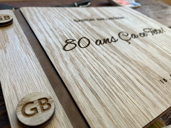 detail of a personalized wooden keepsake album or notebook cover laid flat on a rustic wooden table, with the engraved text  '80 ans Ça ce Fête!', and a personalized name and year