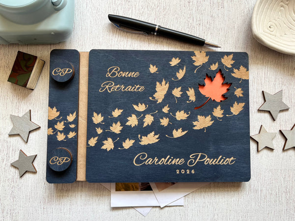 A personalized wooden keepsake notebook dark navy blue with laser-engraved maple leaves, and a small orange cutout revealing an orange first page. The guest book is personalized with the text of your choice and the name of the retiree.  