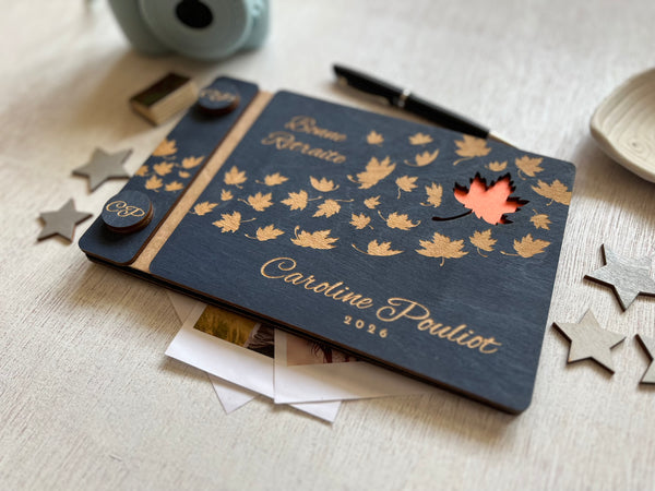 A personalized wooden keepsake notebook dark navy blue with laser-engraved maple leaves, and a small orange cutout revealing an orange first page. The guest book is personalized with the text of your choice and the name of the retiree.  
