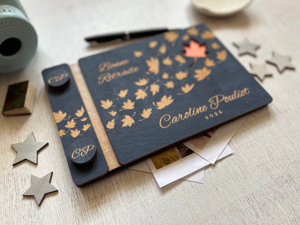 A personalized wooden keepsake notebook dark navy blue with laser-engraved maple leaves, and a small orange cutout revealing an orange first page. The guest book is personalized with the text of your choice and the name of the retiree.  