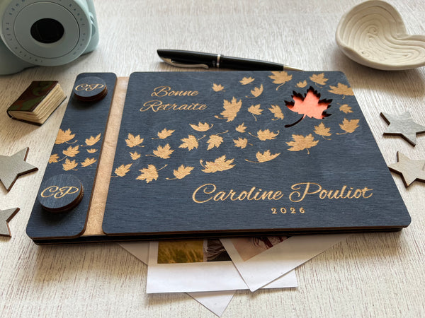 A personalized wooden keepsake notebook dark navy blue with laser-engraved maple leaves, and a small orange cutout revealing an orange first page. The guest book is personalized with the text of your choice and the name of the retiree.  